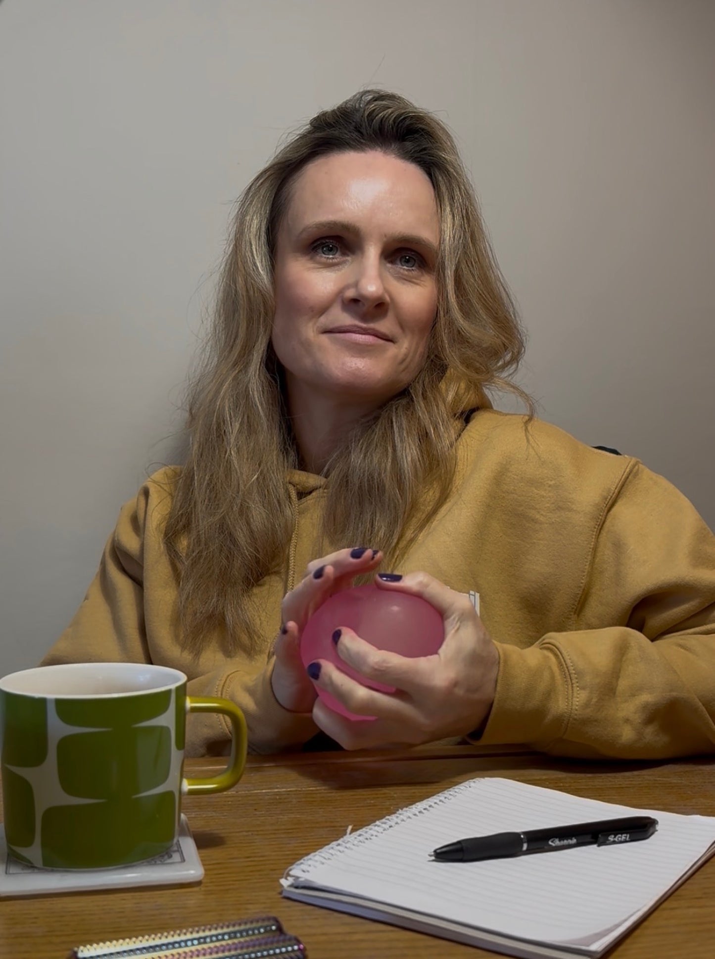 Woman sitting at a table with a pink stress ball, green mug, and notebook.